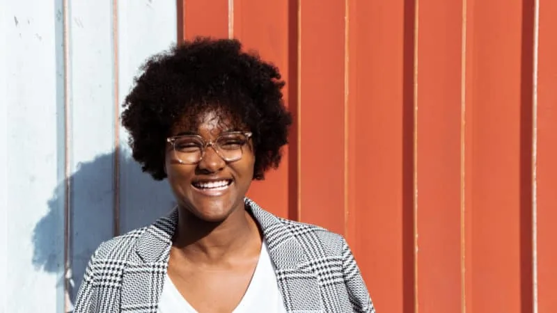 A smiling woman in a plaid jacket standing in front of an orange and white building happy that she is saved
